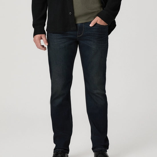 A man wearing dark blue slim-fit jeans, a black cardigan, an olive green shirt, and black leather shoes, standing against a light gray studio background.