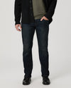 A man wearing dark blue slim-fit jeans, a black cardigan, an olive green shirt, and black leather shoes, standing against a light gray studio background.