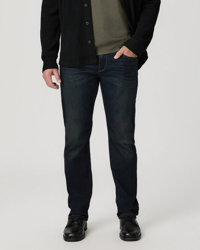 A man wearing dark blue slim-fit jeans, a black cardigan, an olive green shirt, and black leather shoes, standing against a light gray studio background.