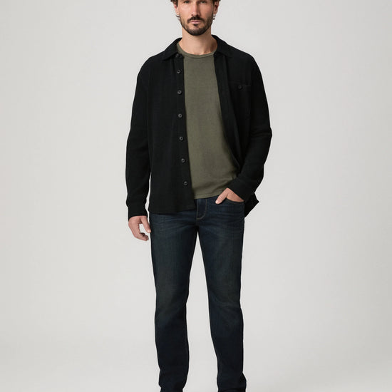 A man modeling a black button-up overshirt over an olive green t-shirt, paired with dark blue jeans and black shoes, against a light gray studio background.
