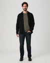 A man modeling a black button-up overshirt over an olive green t-shirt, paired with dark blue jeans and black shoes, against a light gray studio background.