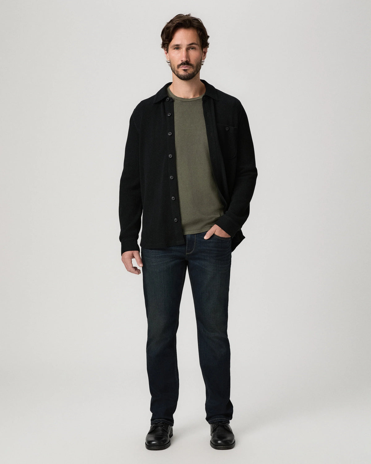 A man modeling a black button-up overshirt over an olive green t-shirt, paired with dark blue jeans and black shoes, against a light gray studio background.
