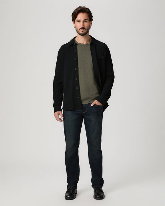 A man modeling a black button-up overshirt over an olive green t-shirt, paired with dark blue jeans and black shoes, against a light gray studio background.