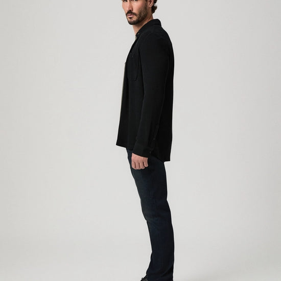 A man in a black blazer, dark jeans, and black leather shoes stands in profile against a light gray studio background.