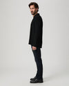 A man in a black blazer, dark jeans, and black leather shoes stands in profile against a light gray studio background.