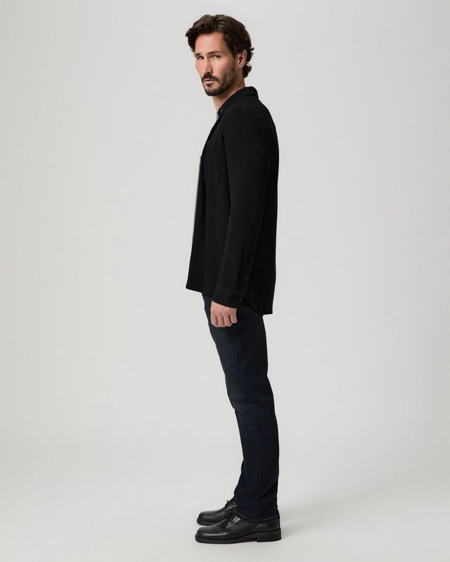 A man in a black blazer, dark jeans, and black leather shoes stands in profile against a light gray studio background.