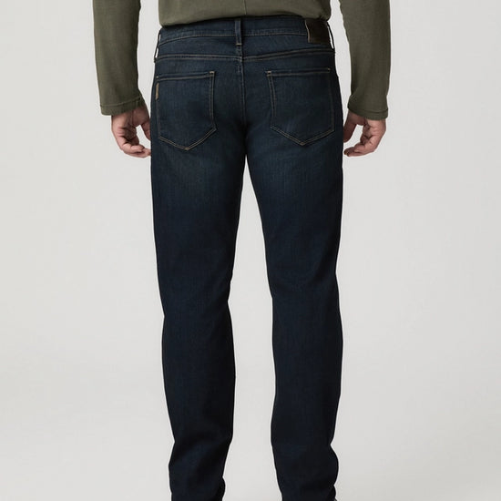 Rear view of a person wearing dark blue denim jeans, an olive green long-sleeve top, and black shoes against a light gray studio background.
