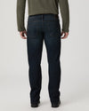 Rear view of a person wearing dark blue denim jeans, an olive green long-sleeve top, and black shoes against a light gray studio background.
