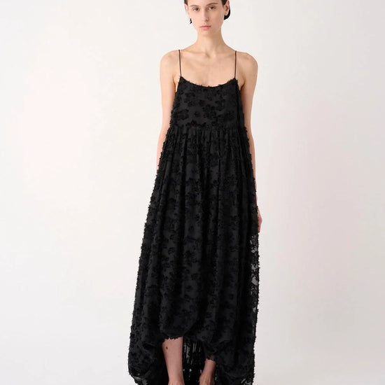 A model stands against a plain light background wearing a long black textured spaghetti-strap dress with a high-low hem and black flat shoes.
