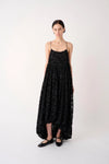 A model stands against a plain light background wearing a long black textured spaghetti-strap dress with a high-low hem and black flat shoes.