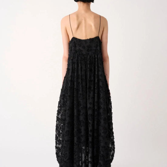 A model viewed from the back wearing a long black spaghetti-strap maxi dress with a textured, floral lace-like pattern and a low open back, set against a plain light gray studio background.
