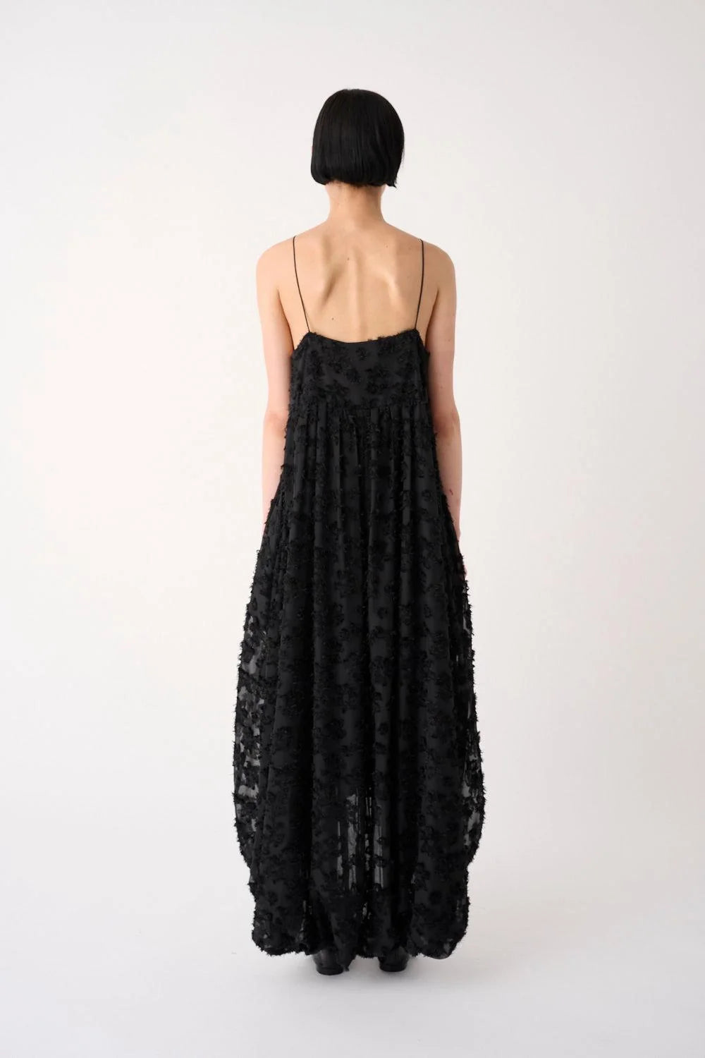 A model viewed from the back wearing a long black spaghetti-strap maxi dress with a textured, floral lace-like pattern and a low open back, set against a plain light gray studio background.