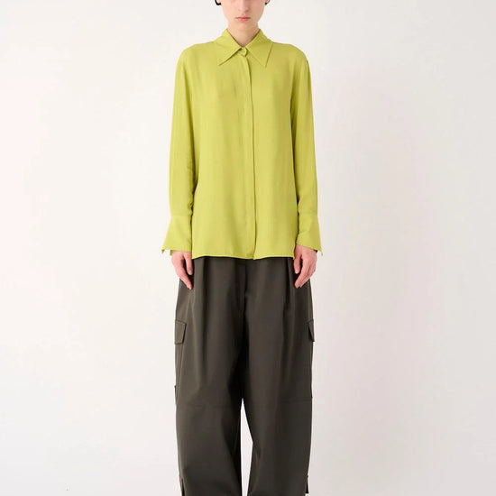 A model stands against a plain light gray studio background wearing a bright yellow-green long-sleeve collared blouse tucked into dark olive wide-leg trousers.