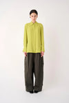 A model stands against a plain light gray studio background wearing a bright yellow-green long-sleeve collared blouse tucked into dark olive wide-leg trousers.