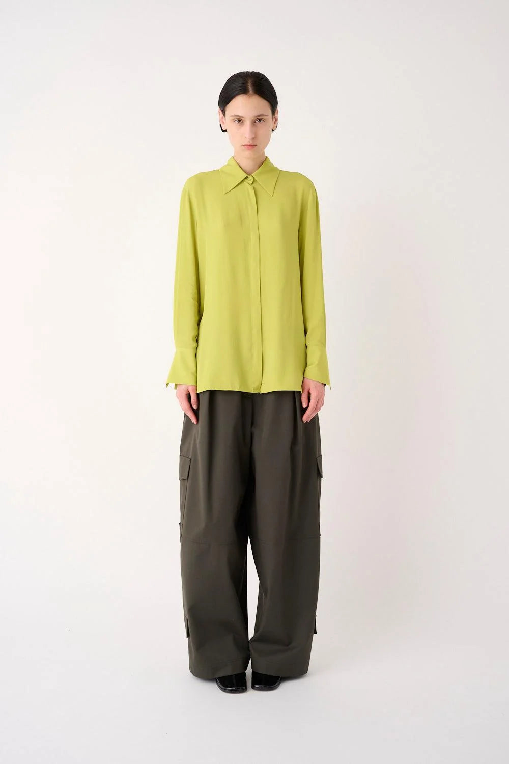 A model stands against a plain light gray studio background wearing a bright yellow-green long-sleeve collared blouse tucked into dark olive wide-leg trousers.