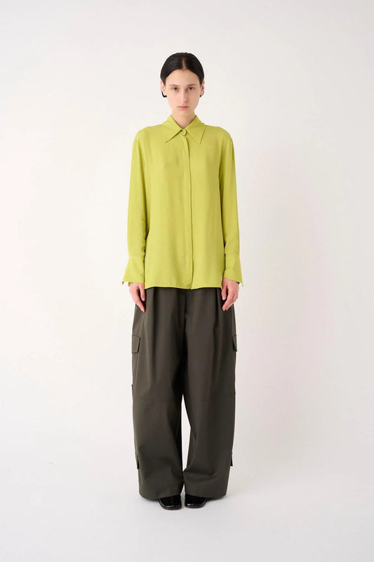 A model stands against a plain light gray studio background wearing a bright yellow-green long-sleeve collared blouse tucked into dark olive wide-leg trousers.