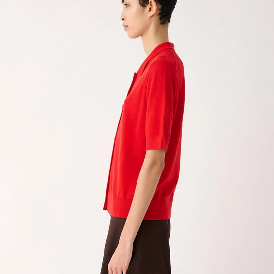 A model shown in profile wearing a bright red short-sleeve button-up shirt and dark brown draped trousers, photographed against a clean light gray studio background.