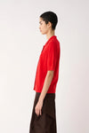 A model shown in profile wearing a bright red short-sleeve button-up shirt and dark brown draped trousers, photographed against a clean light gray studio background.
