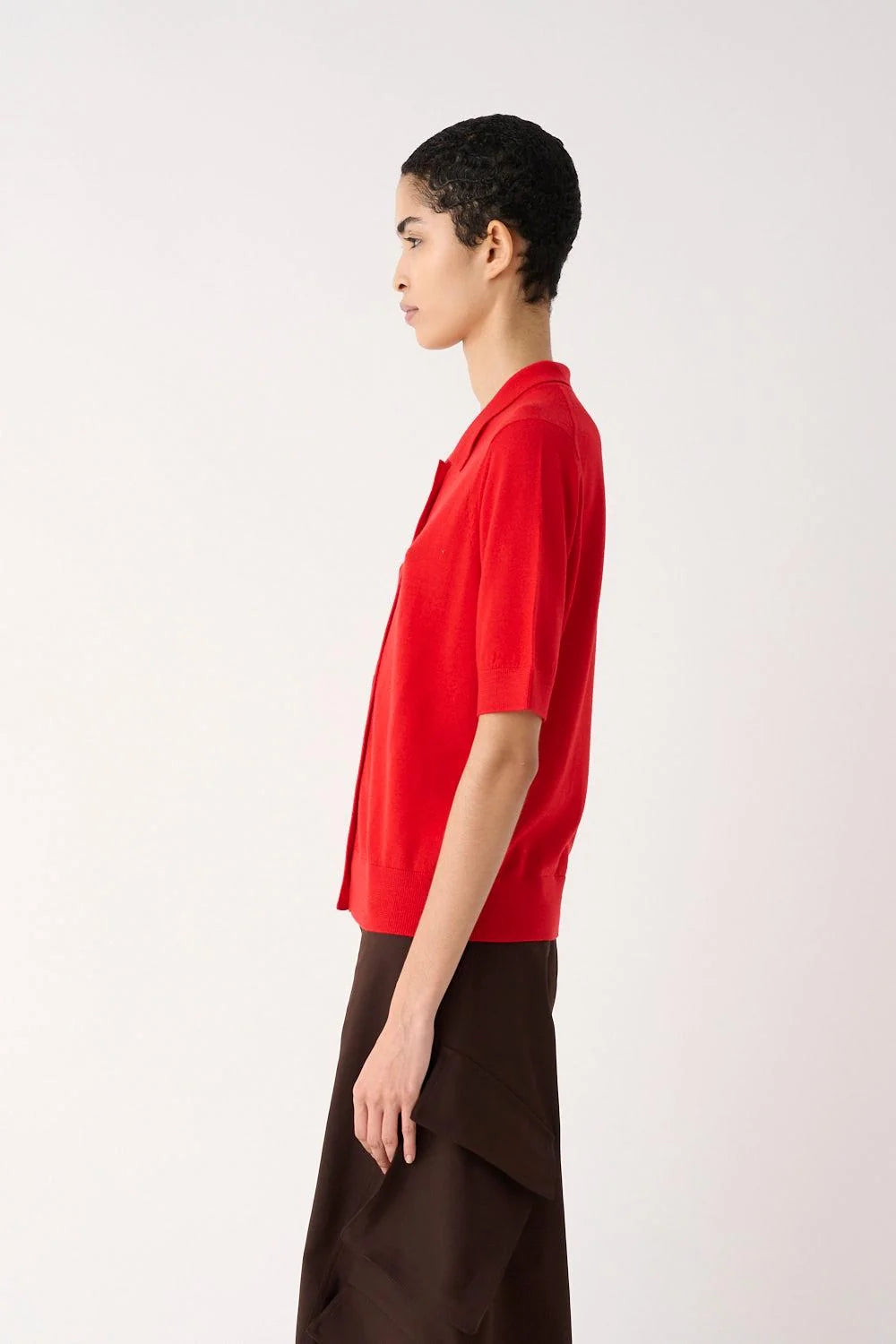 A model shown in profile wearing a bright red short-sleeve button-up shirt and dark brown draped trousers, photographed against a clean light gray studio background.