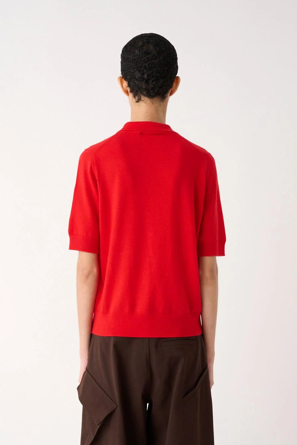 A model shown from the back wearing a bright red short-sleeve knit top and dark brown wide-leg trousers, set against a plain off-white studio background.