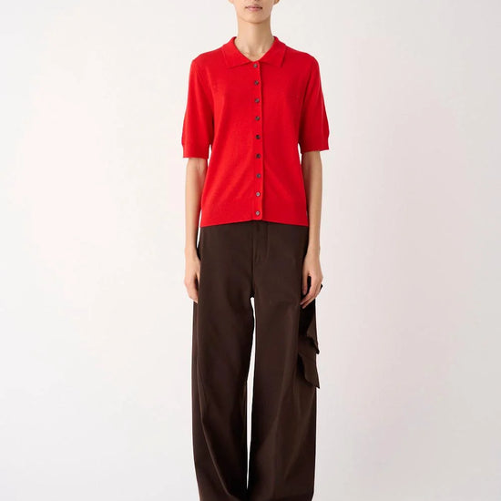 A model stands against a plain light background wearing a bright red short-sleeve button-front top and dark brown wide-leg trousers.