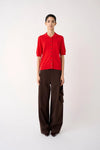 A model stands against a plain light background wearing a bright red short-sleeve button-front top and dark brown wide-leg trousers.