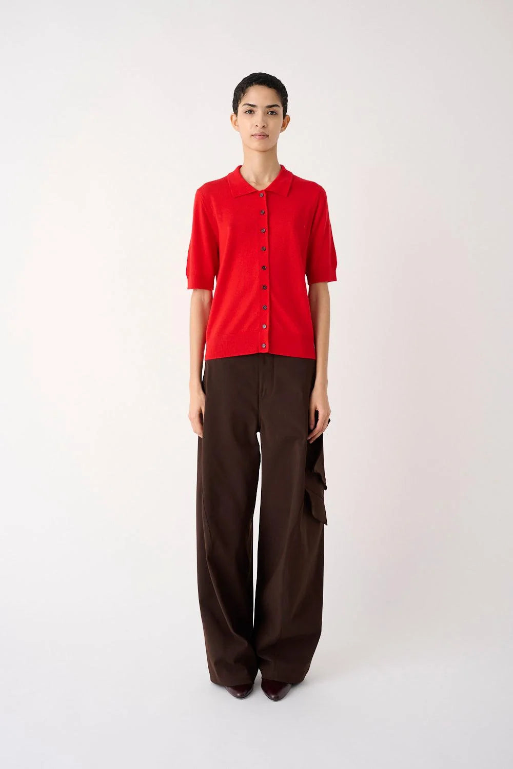 A model stands against a plain light background wearing a bright red short-sleeve button-front top and dark brown wide-leg trousers.