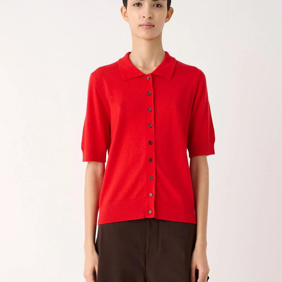 A model wearing a bright red short-sleeve knit polo shirt with dark buttons and dark brown wide-leg trousers, standing against a plain off-white studio background.