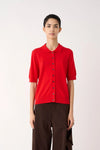 A model wearing a bright red short-sleeve knit polo shirt with dark buttons and dark brown wide-leg trousers, standing against a plain off-white studio background.