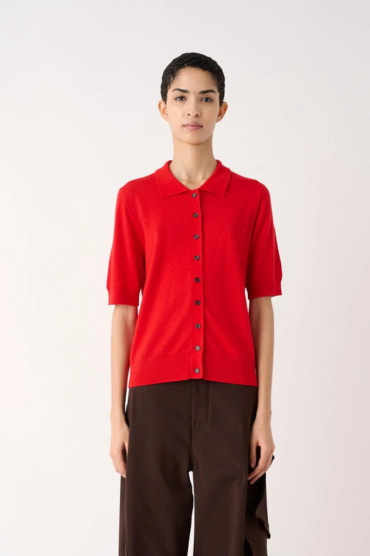 A model wearing a bright red short-sleeve knit polo shirt with dark buttons and dark brown wide-leg trousers, standing against a plain off-white studio background.