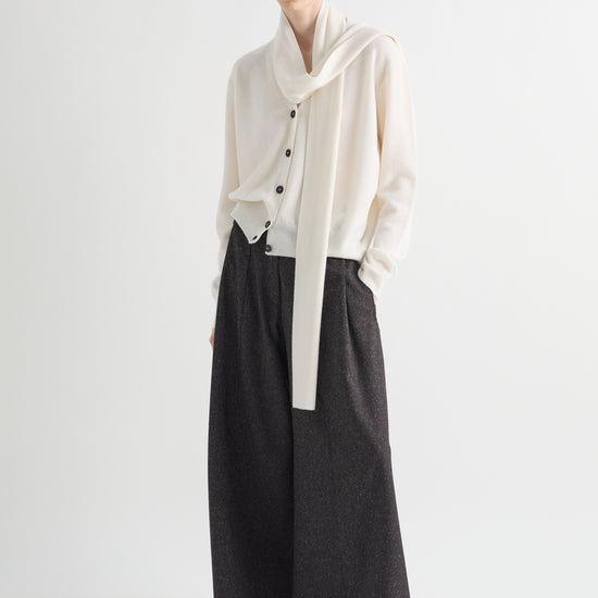 A model wearing a cream-colored draped cardigan with black buttons, paired with wide-leg dark charcoal gray trousers, standing against a plain light gray studio background.