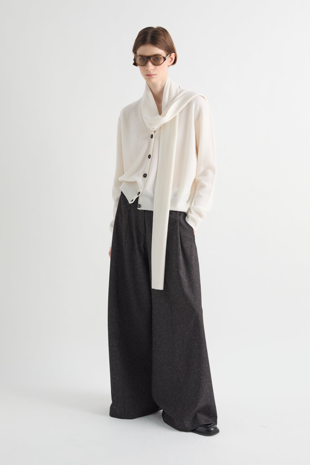 A model wearing a cream-colored draped cardigan with black buttons, paired with wide-leg dark charcoal gray trousers, standing against a plain light gray studio background.