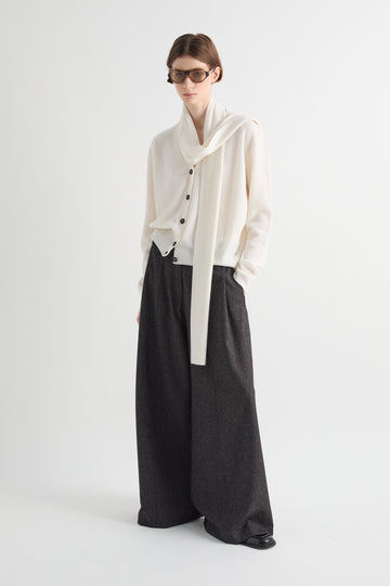 A model wearing a cream-colored draped cardigan with black buttons, paired with wide-leg dark charcoal gray trousers, standing against a plain light gray studio background.