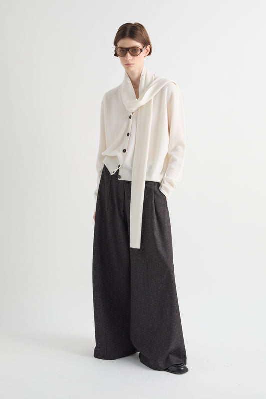 A model wearing a cream-colored draped cardigan with black buttons, paired with wide-leg dark charcoal gray trousers, standing against a plain light gray studio background.