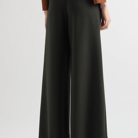 A model shown from the back wearing wide-leg dark green trousers, a brown long-sleeve top, and dark brown heeled sandals against a light gray studio background.