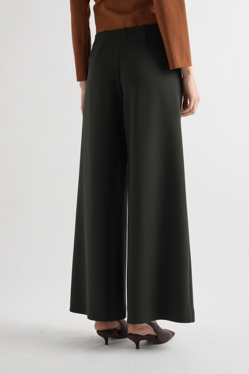 A model shown from the back wearing wide-leg dark green trousers, a brown long-sleeve top, and dark brown heeled sandals against a light gray studio background.