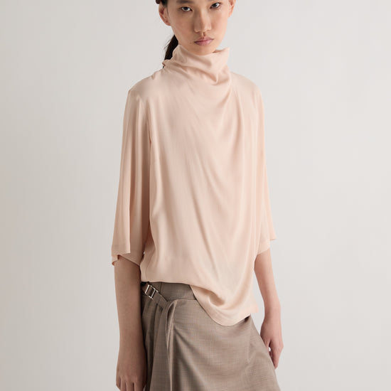 A model wearing a light peach turtleneck top, a brown paperbag skirt, and a dark brown beret, photographed against a neutral light gray studio background.