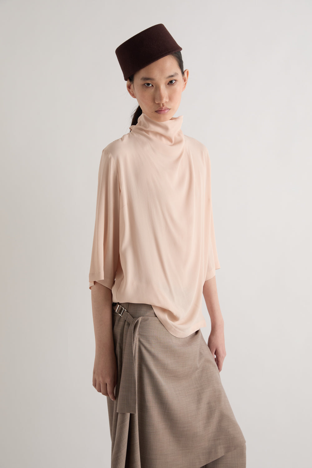 A model wearing a light peach turtleneck top, a brown paperbag skirt, and a dark brown beret, photographed against a neutral light gray studio background.