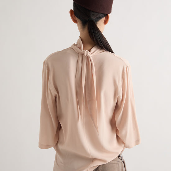 A person shown from the back wearing a pale blush pink long-sleeve blouse with a tie-neck detail, taupe trousers, and a dark brown cap against a plain light gray background.