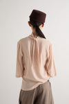 A person shown from the back wearing a pale blush pink long-sleeve blouse with a tie-neck detail, taupe trousers, and a dark brown cap against a plain light gray background.