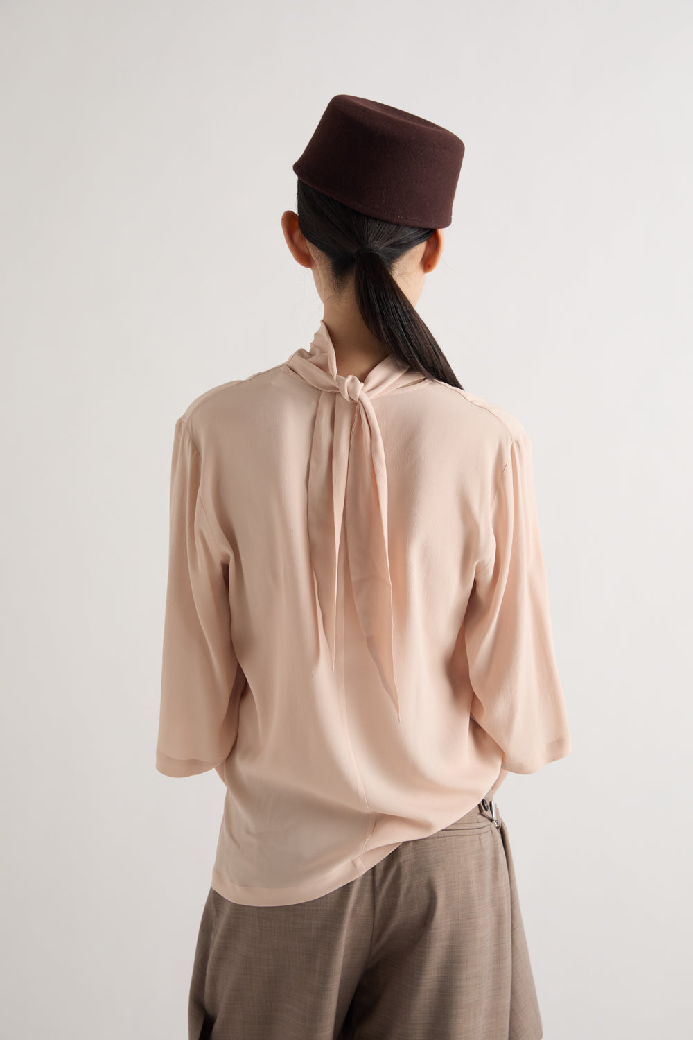 A person shown from the back wearing a pale blush pink long-sleeve blouse with a tie-neck detail, taupe trousers, and a dark brown cap against a plain light gray background.