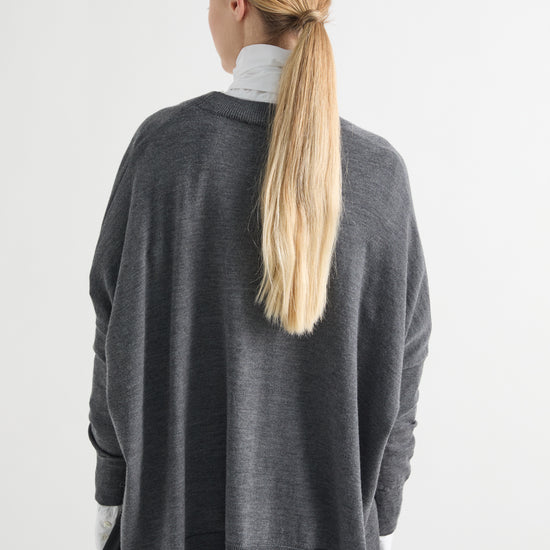 A woman viewed from behind wearing a dark gray oversized knit sweater over a white turtleneck and black pants, against a plain light gray studio background.