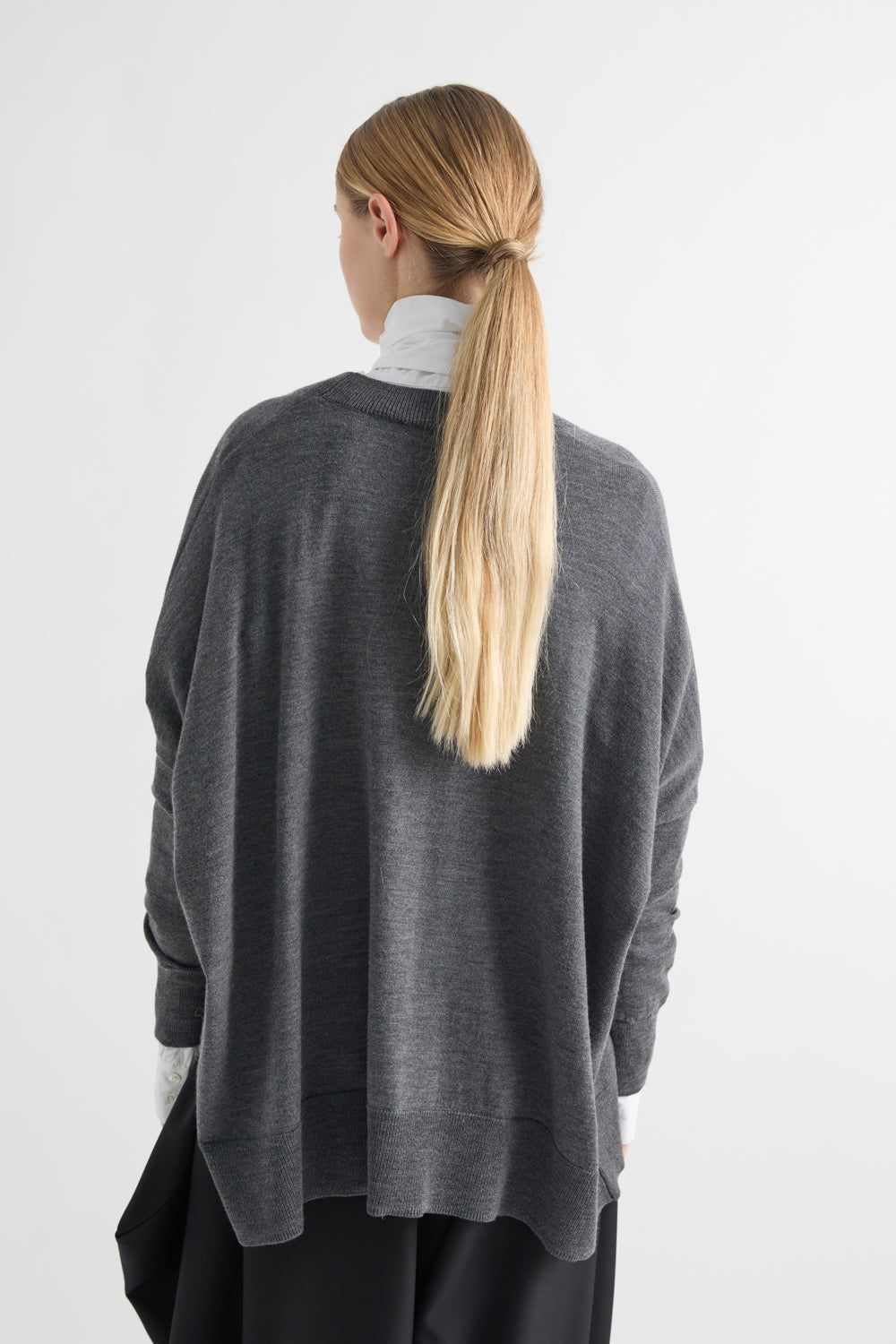 A woman viewed from behind wearing a dark gray oversized knit sweater over a white turtleneck and black pants, against a plain light gray studio background.
