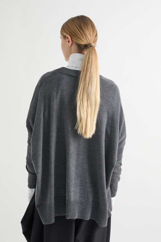 A woman viewed from behind wearing a dark gray oversized knit sweater over a white turtleneck and black pants, against a plain light gray studio background.