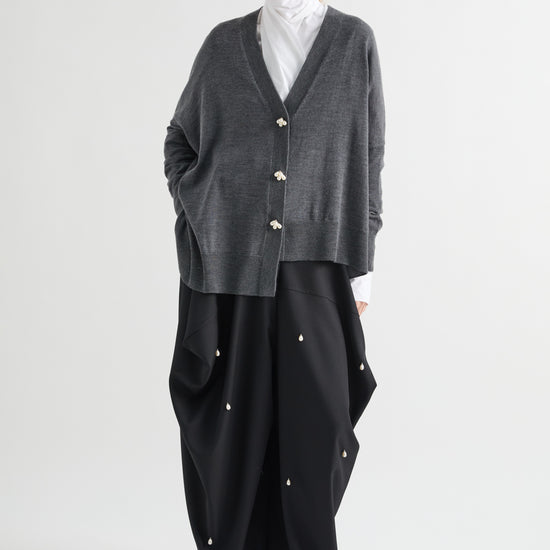 A model stands against a light gray studio background wearing a dark gray knit cardigan with gold-tone buttons over a white turtleneck, paired with loose black wide-leg pants featuring small gold button details.