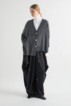A model stands against a light gray studio background wearing a dark gray knit cardigan with gold-tone buttons over a white turtleneck, paired with loose black wide-leg pants featuring small gold button details.