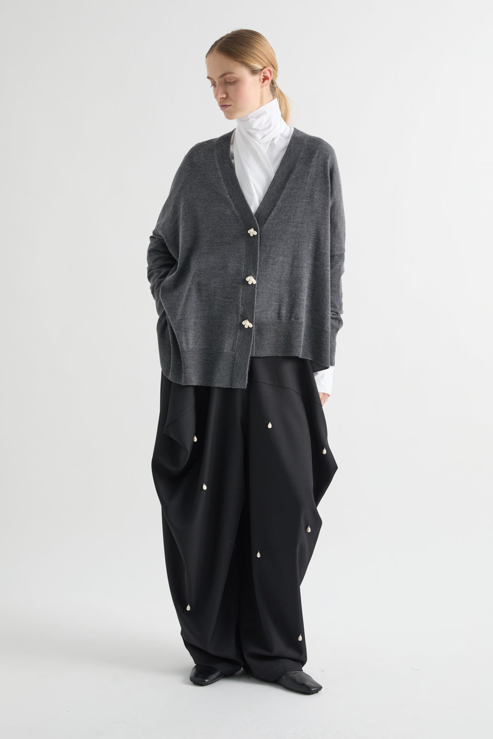 A model stands against a light gray studio background wearing a dark gray knit cardigan with gold-tone buttons over a white turtleneck, paired with loose black wide-leg pants featuring small gold button details.