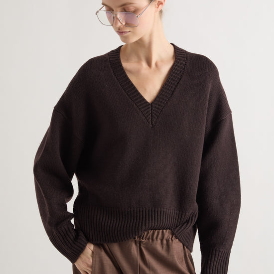 A woman wearing a dark brown V-neck knit sweater and brown trousers, photographed against a plain light gray studio background.