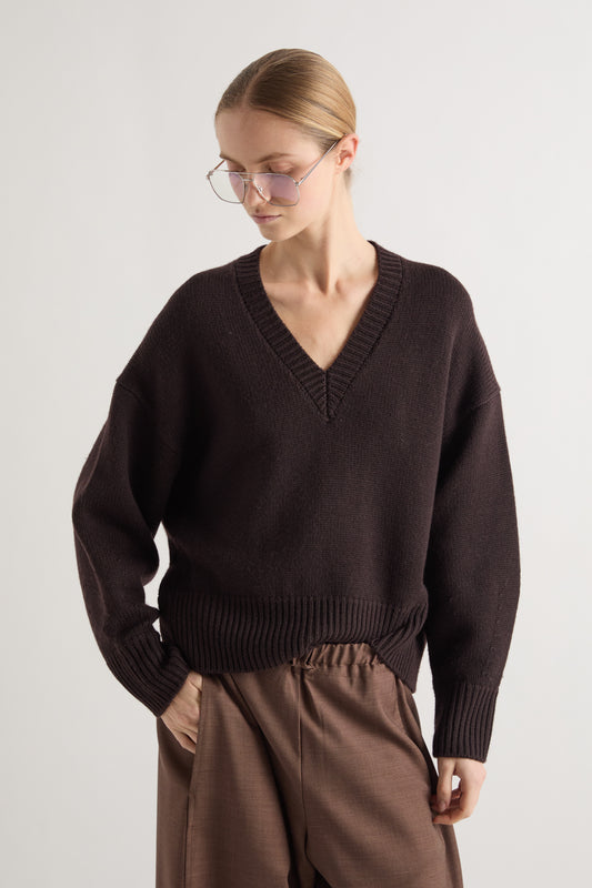 A woman wearing a dark brown V-neck knit sweater and brown trousers, photographed against a plain light gray studio background.