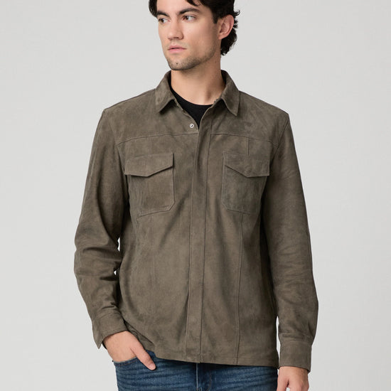 A man wearing a dark brown suede long-sleeve button-up shirt with two chest pockets, paired with blue jeans, photographed against a neutral light gray background.